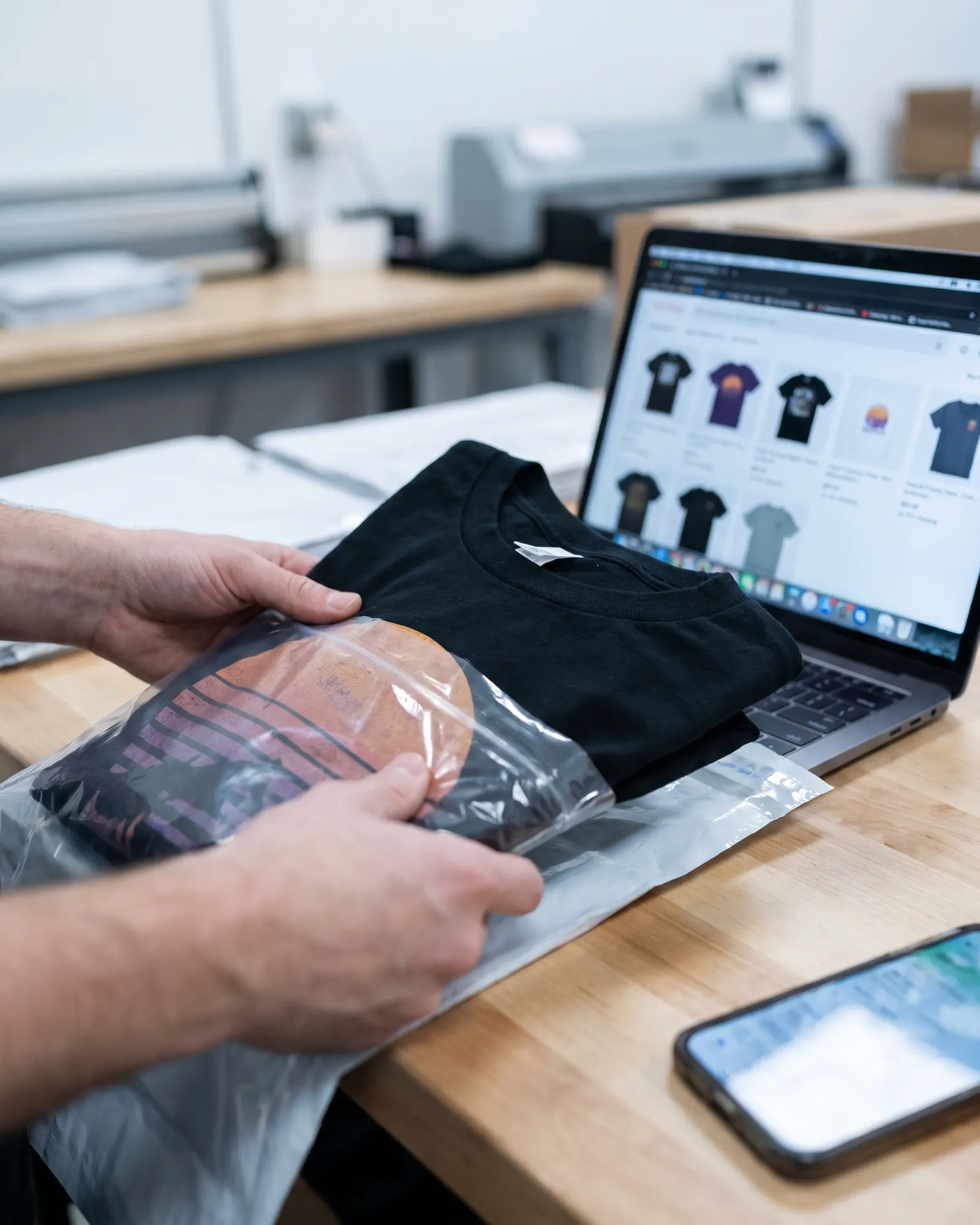 Hands packing a printed tee into a mailer next to a laptop showing an online store.
